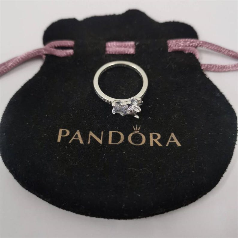 May include: A silver ring with a small rabbit charm, set on a black velvet pouch with a pink drawstring. The pouch has the word "PANDORA" printed in gold. The ring is centered in the image.