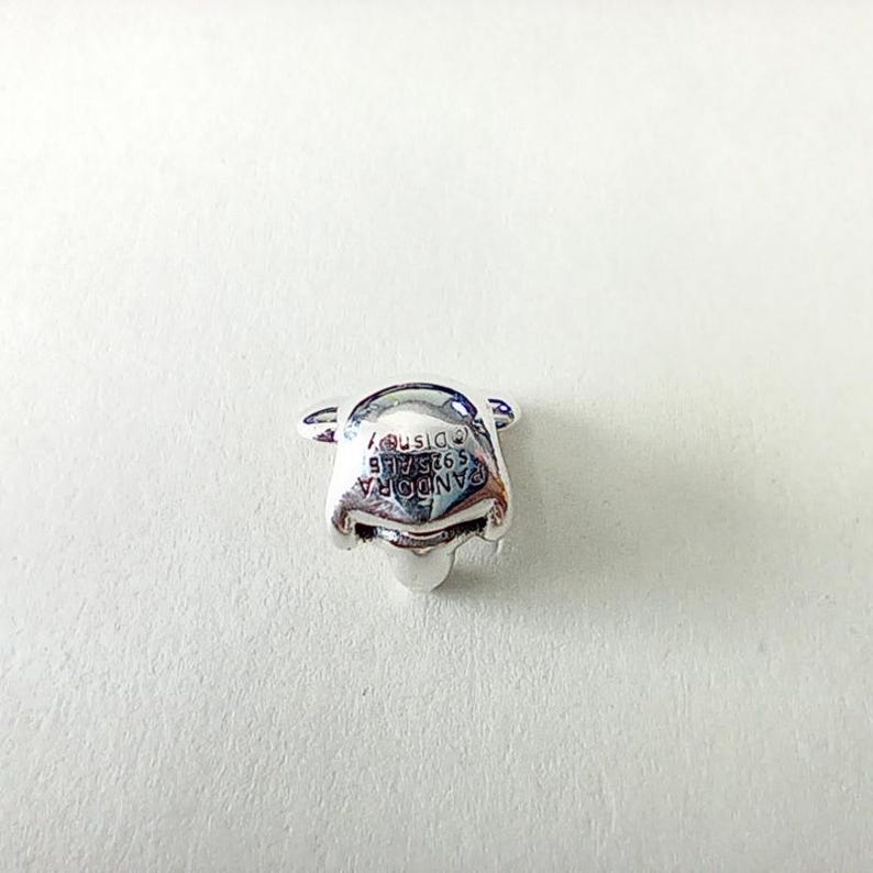 May include: A silver-colored charm in the shape of Winnie the Pooh's face. The charm has the text "PANDORA S925 ALE &copy;Disney" engraved on the front. The charm is on a white background.