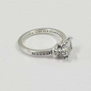 May include: A silver ring with a large, clear gemstone and smaller accent stones. The ring band is engraved with "PANDORA S925 ALE 52". The ring has a detailed, ornate design around the main gemstone, and is set against a white background.