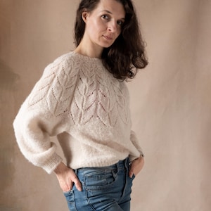 May include: A woman wearing a white knitted sweater with a leaf pattern and blue jeans.