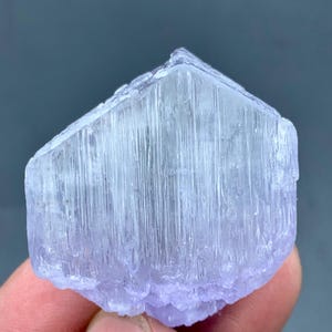 May include: A close-up of a lavender-colored crystal formation. The crystal has a rectangular shape with vertical striations and a slightly rough texture. The color is a pale purple with hints of blue.