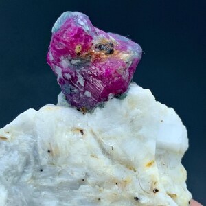 May include: A vibrant, raw ruby gemstone, displaying a deep magenta hue, sits atop a white, textured mineral base. The ruby's crystalline structure is visible, with areas of translucence. The mineral base has a rough, uneven surface.