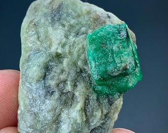 Puede incluir: Un espécimen mineral en bruto que presenta un cristal verde vibrante y translúcido incrustado en una matriz rocosa texturizada de color gris verdoso pálido. El cristal muestra una forma cúbica distintiva con facetas naturales.