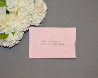 May include: A pink fabric square with the text "Once in a lifetime you meet someone who changes everything" printed on it. The fabric is lying on a gray surface with white carnations in the top left corner.