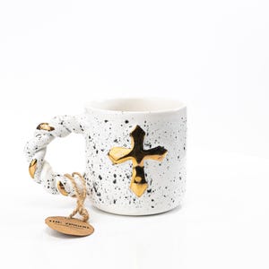 Puede incluir: Taza de cerámica blanca con un esmalte moteado en blanco y negro. La taza tiene un asa de cuerda trenzada y un diseño de cruz dorada. La taza tiene una pequeña etiqueta marrón unida al asa que dice "The Journey".