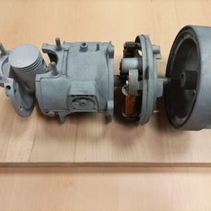 May include: A disassembled silver engine with a piston, cylinder, and fan. The engine is on a wooden surface.