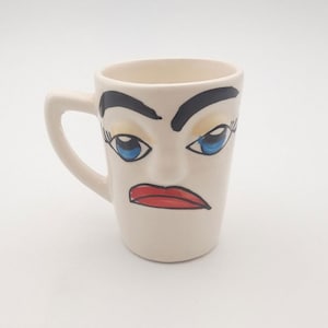 Könnte beinhalten: Weiße Keramiktasse mit handbemaltem Gesicht. Das Gesicht hat blaue Augen, schwarze Augenbrauen und einen roten Mund. Die Tasse hat einen Henkel.