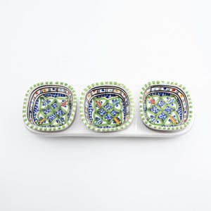 May include: A white ceramic serving dish with three square bowls. Each bowl features a green, blue, and yellow geometric pattern. The rims are decorated with a green dotted border. Suitable for serving appetizers or dips.