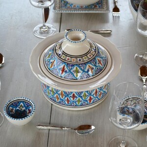 Peut inclure: Un plat de service en céramique blanche avec un couvercle, orné d'un motif géométrique bleu, vert et orange. Le plat est sur une table blanche avec d'autres plats et des couverts.