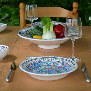 Può includere: Una ciotola da portata in ceramica bianca con un design marocchino colorato, riempita di peperoni verdi, un peperone giallo, un bulbo di finocchio e un peperone rosso. La ciotola è su un tavolo di legno con due bicchieri da vino e una forchetta.
