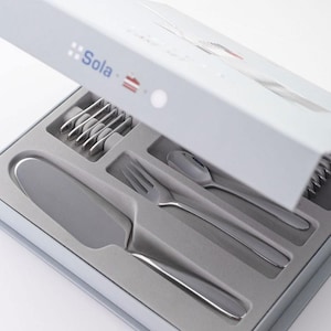 以下が含まれることがあります： グレーと白の箱に入った銀製の食器セット。食器には、ナイフ、フォーク、スプーン、ケーキサーバーが含まれています。箱には「Sola」という文字が印刷されています。