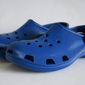 Könnte beinhalten: Ein Paar blaue Clogs mit mehreren Belüftungslöchern und einem schwenkbaren Fersenriemen. Die Schuhe bestehen aus einem festen, geformten Material und sind für den Freizeitgebrauch konzipiert. Die Clogs werden vor weißem Hintergrund gezeigt.