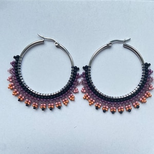 Puede incluir: Un par de pendientes de aro plateados con flecos de cuentas. Los flecos presentan un degradado de colores, incluyendo negro, morado, rosa y naranja. Los pendientes están diseñados con un patrón de cuentas.