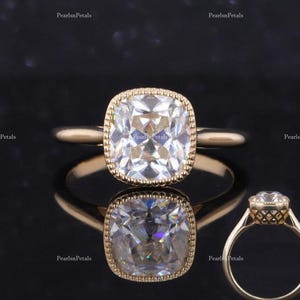 May include: A gold ring with a large, square-shaped, clear gemstone set in a bezel setting. The gemstone has a cushion cut and is surrounded by a delicate, beaded border. The ring is shown on a black background.