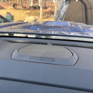 Può includere: Vista ravvicinata di una griglia di altoparlante nera sul cruscotto di un'auto. L'altoparlante è rettangolare e ha un piccolo pulsante nero con un simbolo triangolare al centro.