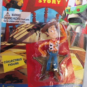 May include: A Woody collectible figure from Toy Story in original packaging. The plastic-encased figure wears a brown cowboy hat, yellow and red plaid shirt, cow-print vest, and blue pants. The backing card features Buzz Lightyear and text.