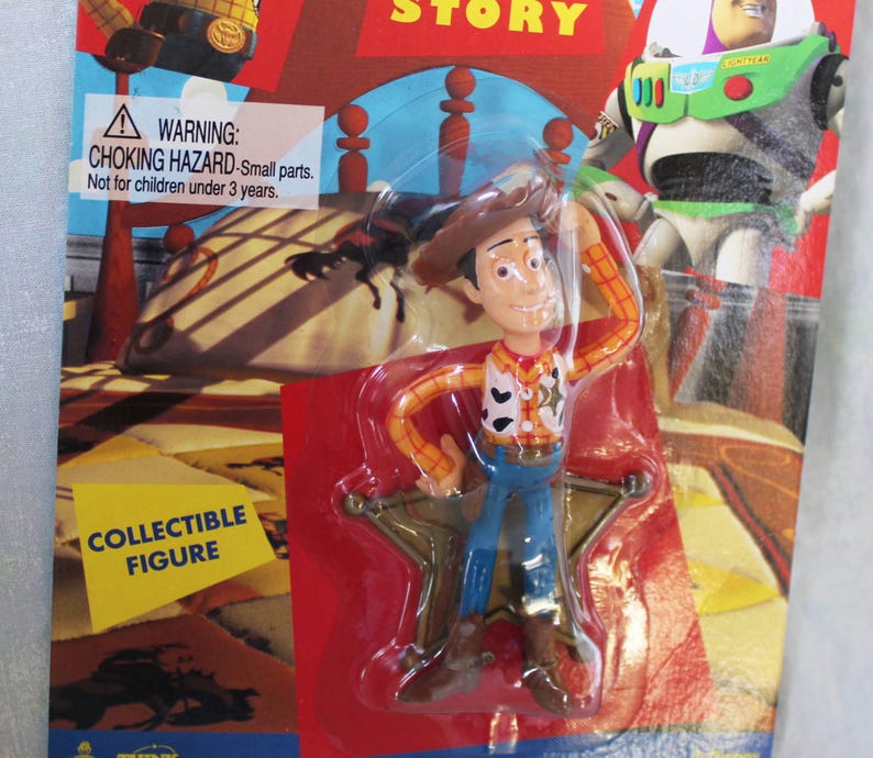 May include: A Woody collectible figure from Toy Story in original packaging. The plastic-encased figure wears a brown cowboy hat, yellow and red plaid shirt, cow-print vest, and blue pants. The backing card features Buzz Lightyear and text.