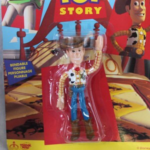 May include: A bendable Woody action figure from Toy Story, packaged in a clear plastic blister on a cardboard backing. The figure wears a yellow plaid shirt, cow-print vest, blue pants, brown boots, and a brown cowboy hat, with one arm raised.