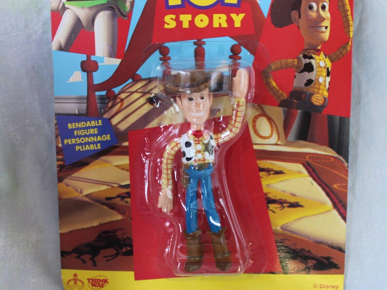 May include: A bendable Woody action figure from Toy Story, packaged in a clear plastic blister on a cardboard backing. The figure wears a yellow plaid shirt, cow-print vest, blue pants, brown boots, and a brown cowboy hat, with one arm raised.