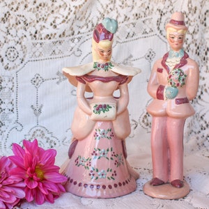 May include: A pair of vintage ceramic figurines, a woman and a man, in pastel pink and cream colors. The woman wears a dress with floral accents and holds a small bag. The man wears a suit and holds a bouquet. The figurines are approximately 8 inches tall.