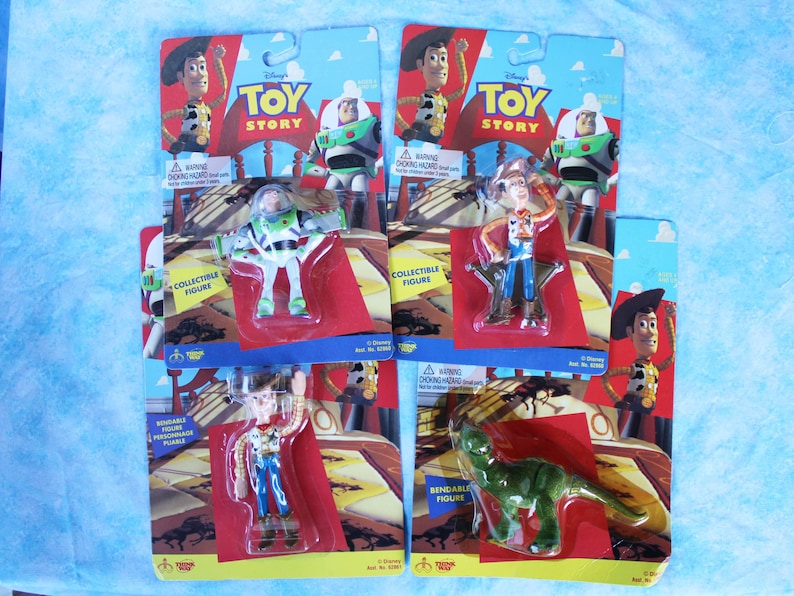 May include: Four Disney Toy Story collectible figures in original blister packaging. The set includes two Woody figures, one Buzz Lightyear figure, and one green dinosaur figure. Each card features character artwork and the Toy Story logo.