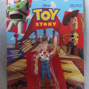 May include: A bendable Woody action figure from Toy Story in its original packaging. The card features Buzz Lightyear and Woody graphics, with the text Toy Story, Ages 3 and Up, and Bendable Figure. The figure wears a cowboy hat, vest, and boots.