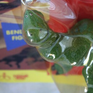 May include: A green textured lizard figurine encased in clear molded plastic packaging. The lizard has two small white eyes on its head. Behind the plastic, a portion of a cardboard box is visible with the partial text BENDY FIG.