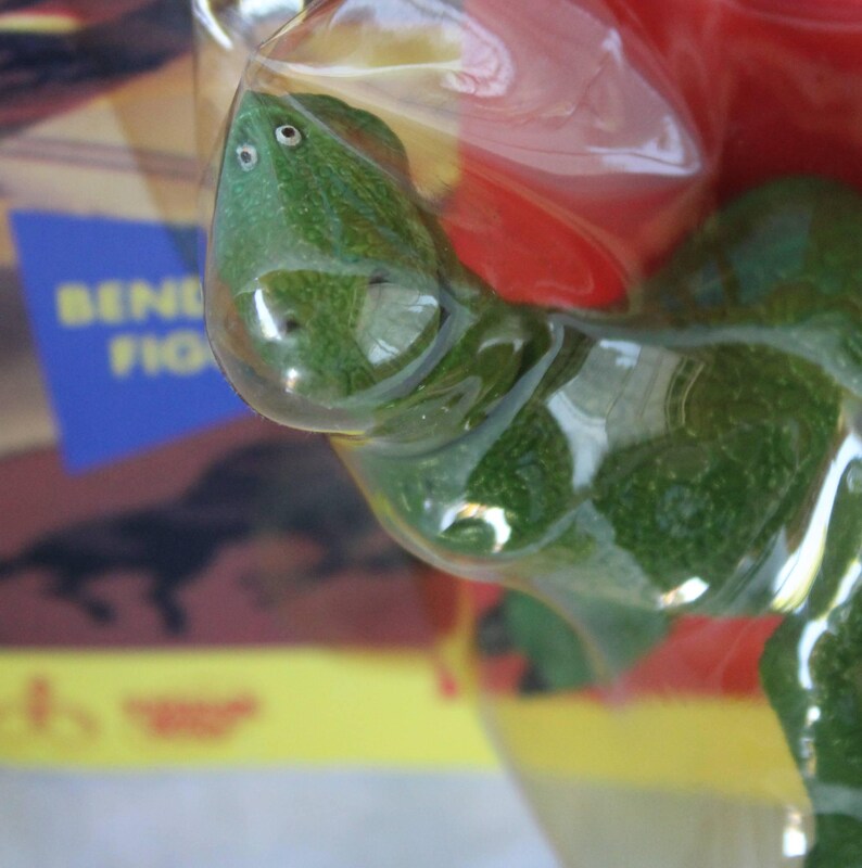 May include: A green textured lizard figurine encased in clear molded plastic packaging. The lizard has two small white eyes on its head. Behind the plastic, a portion of a cardboard box is visible with the partial text BENDY FIG.