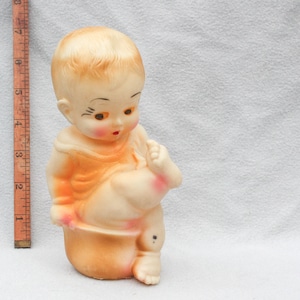 May include: Vintage rubber baby doll toy, featuring a seated pose with one leg raised. The doll has painted facial features, including rosy cheeks and a red mouth. The doll is approximately 5 inches tall.