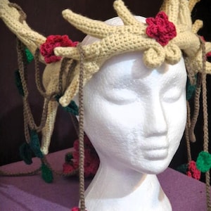 May include: A crocheted crown with antlers, leaves, and red flowers. The crown is worn on a white mannequin head.