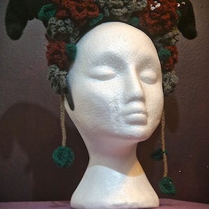 May include: A crocheted headband with black horns, red and green flowers, and dangling green pom-poms. The headband is displayed on a white mannequin head.