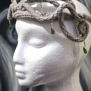 May include: A grey crocheted crown with green and clear gemstone accents. The crown is worn on a white mannequin head.