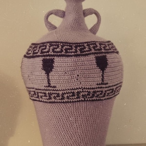 Könnte beinhalten: Eine lavendelfarbene gehäkelte Vase mit zwei Henkeln und einem schmalen Hals. Die Vase hat einen schwarzen geometrischen Rand und zwei schwarze Weinglas-Silhouetten. Die handgefertigte Vase ist ein Dekorationsartikel.