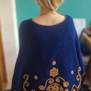 May include: A handmade, navy blue crochet poncho with a round neckline and decorative tan-coloured floral and swirl patterns along the bottom edge. The poncho is a wearable accessory.