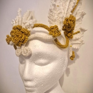 May include: A handmade crochet headpiece in white and gold, displayed on a mannequin head. The headpiece features white crochet leaves and gold crochet flowers and accents. The headpiece is designed to be worn on the head.