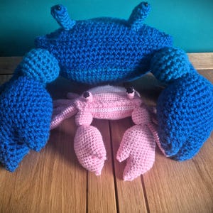 Puede incluir: Dos cangrejos de crochet, uno azul y otro rosa, están sentados sobre una superficie de madera. El cangrejo azul es más grande y tiene un diseño más detallado. El cangrejo rosa es más pequeño y tiene un diseño más simple.