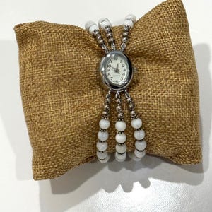 May include: A silver watch with a white face and white bead bracelet. The watch is on a brown hessian cushion.