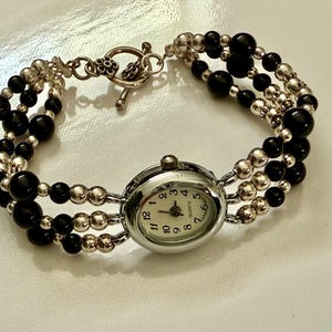 May include: A silver watch with a white face and black numbers. The watch is attached to a bracelet made of black and silver beads.