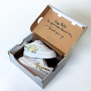 May include: White sneakers with gold accents and floral designs inside a gray box. The box lid has handwritten text that reads "Say hello! to your new favorite Thank you". The sneakers have the word "Σταμάτη" written on the side.