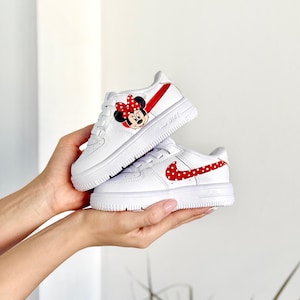 May include: White toddler sneakers with red accents. One shoe features a Minnie Mouse graphic and the other has a red polka dot Nike swoosh. The shoes are held in a person's hands.