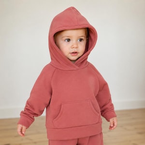 May include: A dusty rose-colored hooded sweatshirt with a kangaroo pocket. The sweatshirt is part of a matching set, and the hood is up. The outfit is made of a soft material.