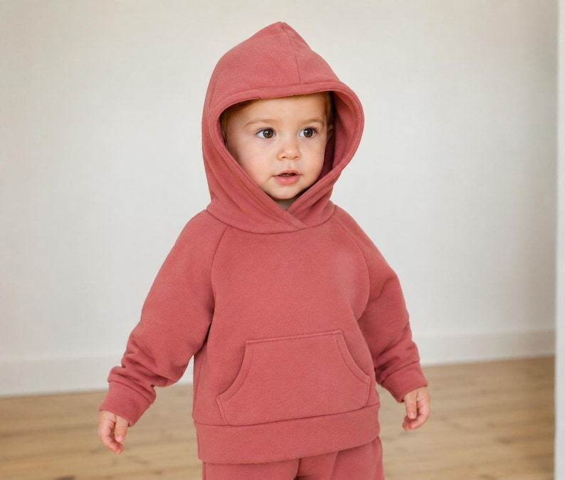 May include: A dusty rose-colored hooded sweatshirt with a kangaroo pocket. The sweatshirt is part of a matching set, and the hood is up. The outfit is made of a soft material.
