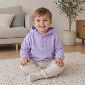 May include: A young child wearing a light purple hoodie with wooden buttons and beige pants sits on a white rug. The child is smiling. A beige sofa with pillows is in the background.