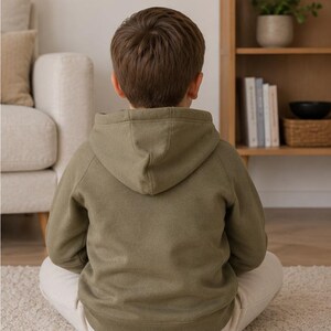 May include: A person wearing an olive green hoodie with a hood, sitting cross-legged on a white rug. The person is facing away from the camera. The background includes a beige sofa and a wooden bookshelf.