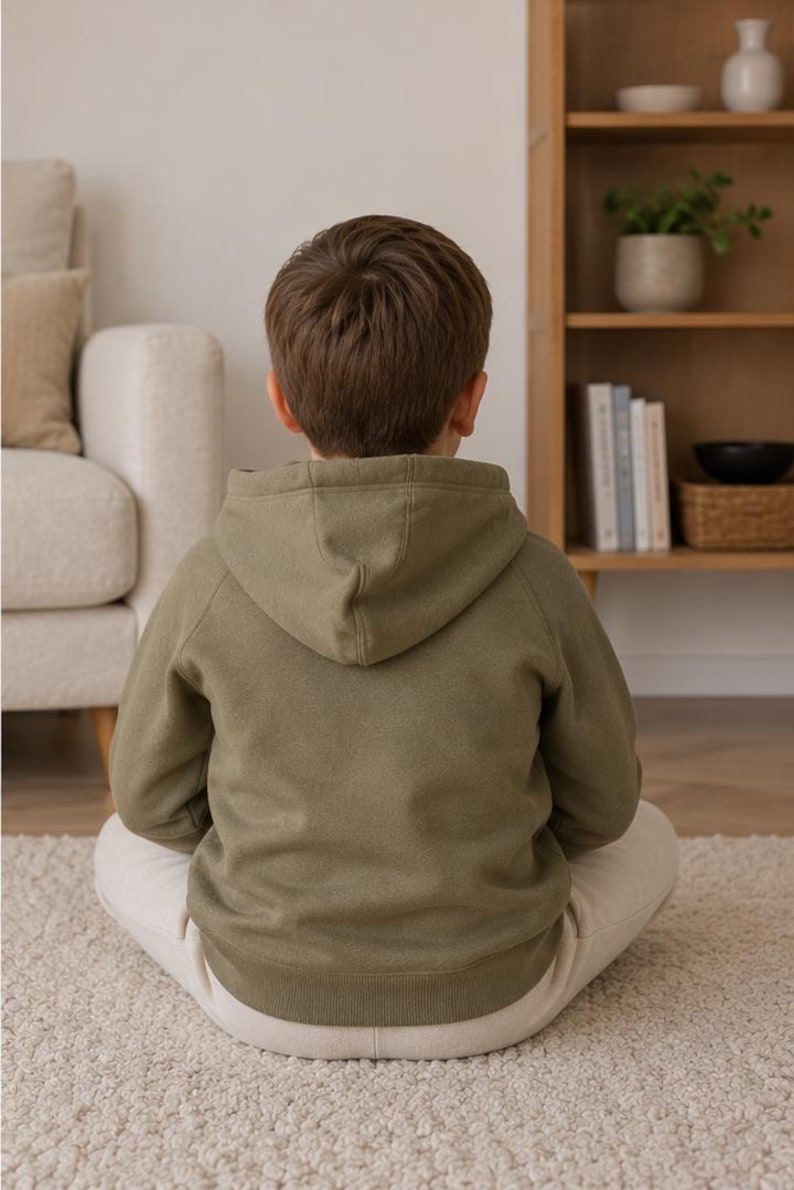 May include: A person wearing an olive green hoodie with a hood, sitting cross-legged on a white rug. The person is facing away from the camera. The background includes a beige sofa and a wooden bookshelf.