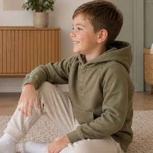 May include: An olive green hooded sweatshirt with a three-button placket and a front pocket. The sweatshirt is paired with cream-colored sweatpants. The person is sitting on a beige carpet.