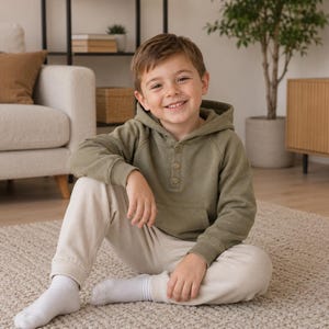 May include: A young person wearing a green hooded sweatshirt and beige sweatpants sits on a beige rug. The person is smiling and wearing white socks. A beige sofa and wooden cabinet are in the background.