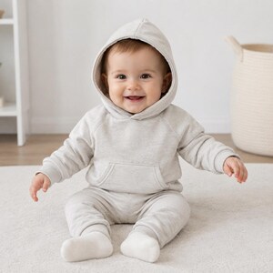 May include: A smiling baby wearing a light gray hoodie and matching sweatpants. The baby is sitting on a white rug. The outfit has a kangaroo pocket on the front of the hoodie.