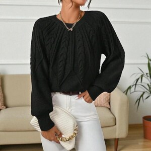 May include: A black cable-knit sweater with a round neckline and long sleeves. The sweater has a textured pattern and ribbed cuffs. The person is wearing white pants and holding a white clutch with a gold chain strap. A gold necklace is also visible.