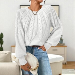 May include: A white cable-knit sweater with a round neckline and long, puffy sleeves. The sweater has a textured pattern on the front and sleeves. The model is wearing blue jeans and holding a white clutch with a gold chain.
